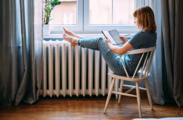 Femme lisant un livre, pieds sur un radiateur : économies de chauffage